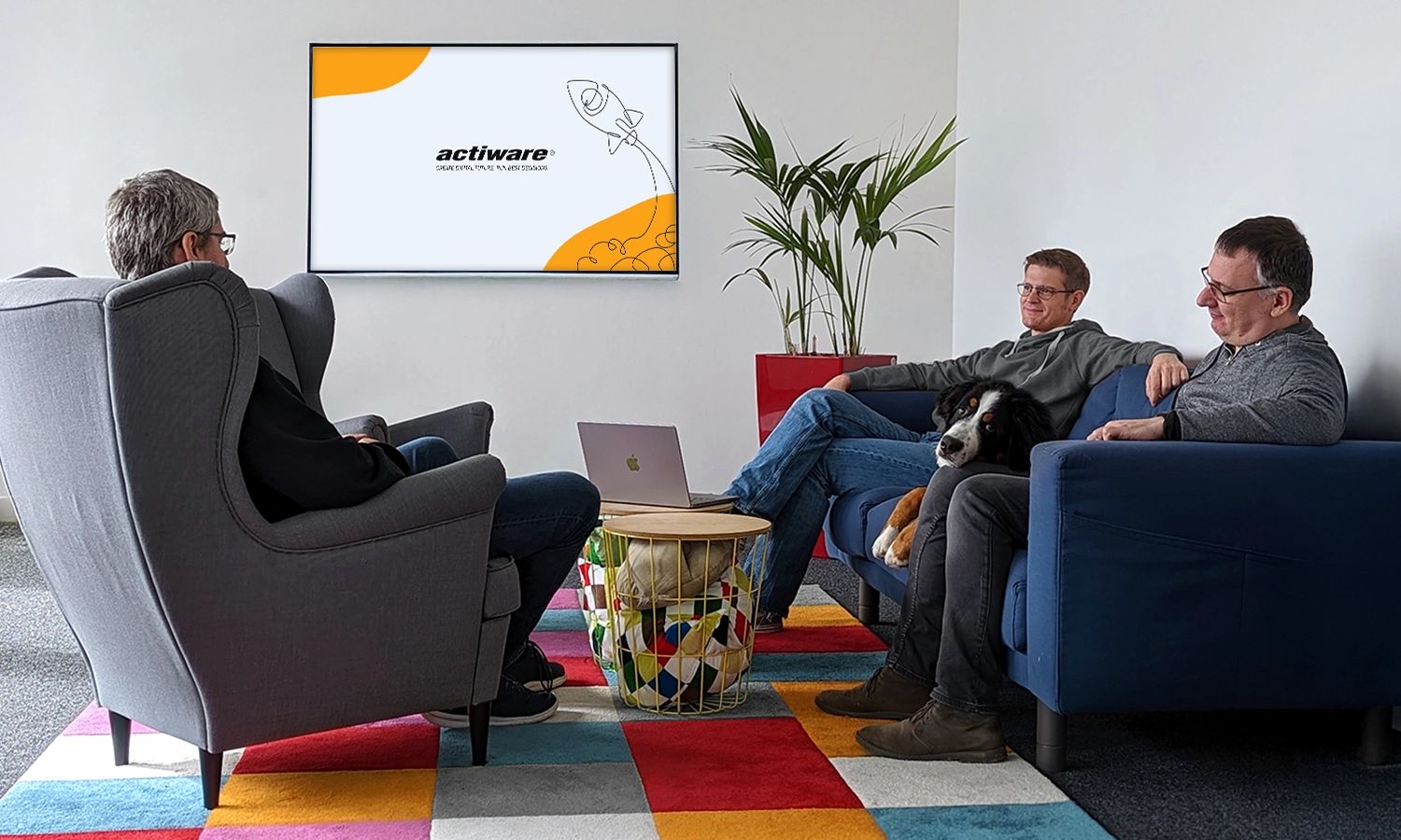 Drei Männer und ein Hund sitzen entspannt im Büro-Loungebereich vor einem Bildschirm mit ACTIWARE-Logo.