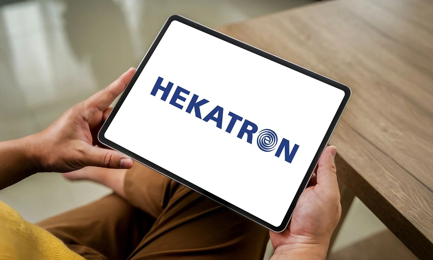 Person hält ein Tablet, auf dessen Bildschirm das Logo von HEKATRON mit blauem Schriftzug und stilisiertem Spiralmotiv im Buchstaben O angezeigt wird.